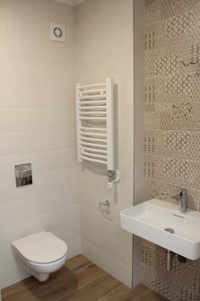 A modern bathroom featuring beige tiles on the walls and patterned tiles on one side. The floor is covered with wood-like tiles. There is a wall-mounted toilet with a silver flush button. Next to it, a white wall-mounted towel radiator is present. A white rectangular ceramic sink is fixed on the wall with a modern faucet.