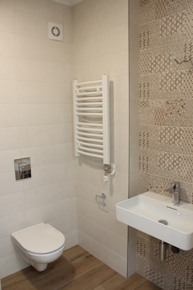 A modern bathroom featuring beige tiles on the walls and patterned tiles on one side. The floor is covered with wood-like tiles. There is a wall-mounted toilet with a silver flush button. Next to it, a white wall-mounted towel radiator is present. A white rectangular ceramic sink is fixed on the wall with a modern faucet.