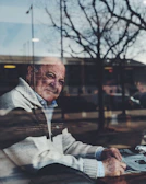 An elderly man sitting quietly by a window, looking out over a city scarred by conflict.