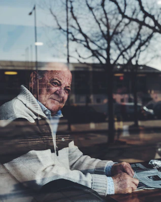 An elderly man sitting quietly by a window, looking out over a city scarred by conflict.