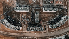 Aerial view of a residential area featuring multiple large apartment buildings arranged in a grid-like pattern. Cars are parked along winding roads, and pathways connect the buildings with patches of grass and bare land visible throughout. Roads and infrastructure border the residential complex, with snow patches indicating a cold season.
