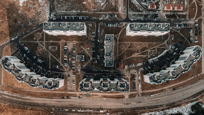 Aerial view of a residential area featuring multiple large apartment buildings arranged in a grid-like pattern. Cars are parked along winding roads, and pathways connect the buildings with patches of grass and bare land visible throughout. Roads and infrastructure border the residential complex, with snow patches indicating a cold season.
