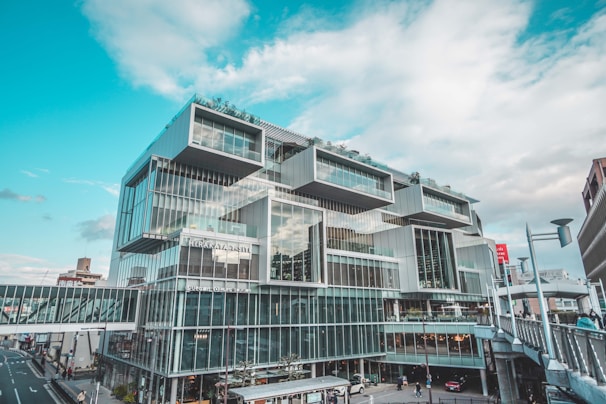 A modern, multi-story building with a modular and geometric architectural design. It features large glass windows and panels, creating a sleek and futuristic appearance. There are people walking on a skybridge connecting to the building, and a cloudy sky in the background.