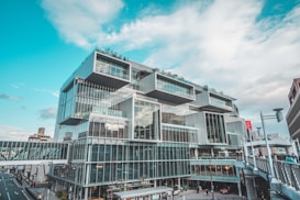 A modern, multi-story building with a modular and geometric architectural design. It features large glass windows and panels, creating a sleek and futuristic appearance. There are people walking on a skybridge connecting to the building, and a cloudy sky in the background.