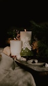A cozy and warm setting featuring two lit candles placed amidst pine cones and greenery. A decorative piece resembling a small Christmas tree with a star on top is present, adding a festive touch. Soft textures like fabric and a wooden surface create a comforting atmosphere.