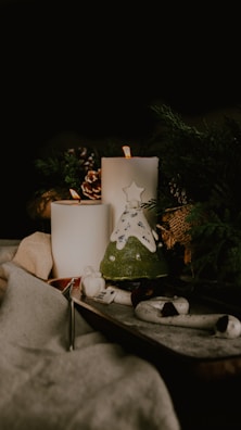 A cozy and warm setting featuring two lit candles placed amidst pine cones and greenery. A decorative piece resembling a small Christmas tree with a star on top is present, adding a festive touch. Soft textures like fabric and a wooden surface create a comforting atmosphere.