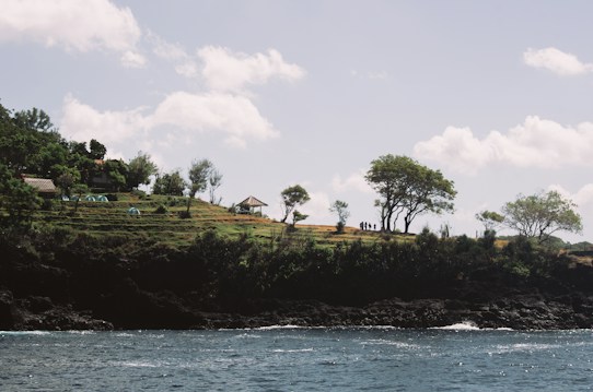 A serene coastal landscape with gentle waves meeting a rocky shoreline. Above the shoreline, lush greenery covers small hills where tents and a pavilion are visible. Trees are scattered across the hills, and figures are seen walking along a path. The sky is clear with some fluffy clouds.