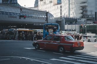 A bustling urban scene featuring a red taxi crossing an intersection. In the background, a crowd of people waits at a bus stop. Several buses, including a blue one with cartoon graphics, are nearby. The street is lined with tall buildings, some under construction.
