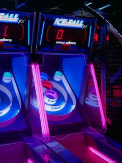Brightly lit arcade skee-ball machines with neon pink and blue lights dominate the scene. The game displays and scoring areas are glowing under the vibrant lighting, creating an enticing atmosphere.