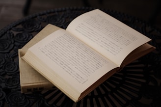 An open book with text in Japanese is placed on an intricately designed black metal table. The pages are slightly yellowed, indicative of age or classic literature. The table has a floral pattern, adding an artistic element to the scene.