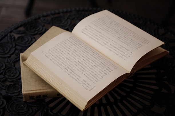 An open book with text in Japanese is placed on an intricately designed black metal table. The pages are slightly yellowed, indicative of age or classic literature. The table has a floral pattern, adding an artistic element to the scene.