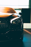 A close-up of a sleek, modern car with sharp headlights and a glossy finish. The vehicle is positioned indoors with soft lighting highlighting its contours. The photo emphasizes the car's luxurious design and high-quality details.