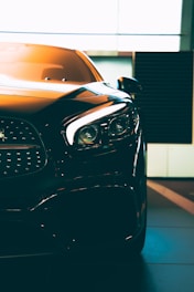 A close-up of a sleek, modern car with sharp headlights and a glossy finish. The vehicle is positioned indoors with soft lighting highlighting its contours. The photo emphasizes the car's luxurious design and high-quality details.