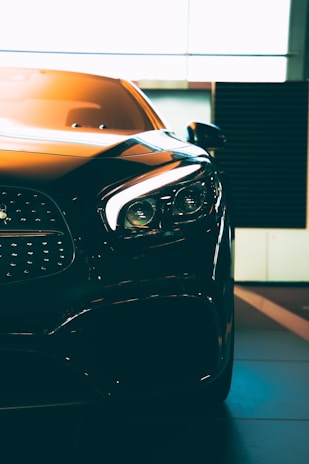 A close-up of a sleek, modern car with sharp headlights and a glossy finish. The vehicle is positioned indoors with soft lighting highlighting its contours. The photo emphasizes the car's luxurious design and high-quality details.