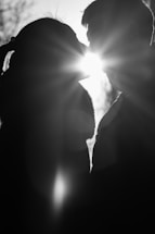 A black and white silhouette of two people kissing with the sun shining brightly between them, creating a halo effect and lens flare. The scene is outdoors, possibly during sunrise or sunset, with blurred background details.