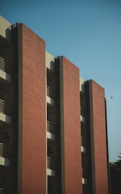 A multi-story building with a modern design featuring several vertical sections of red brick walls. Each segment is divided by concrete columns, and the structure includes balconies with metal railings. The sky is clear, contributing to a clean and minimalistic appearance.