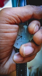 A hand tightly grips a wristwatch, with focus on the watch's face displaying clear numbers and text indicating it is a quartz water-resistant watch. The strap is made of black leather. The lighting highlights the texture of the skin and the smooth surface of the watch.