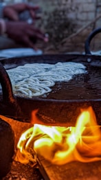 A traditional cooking setup with a large metal pan over an open flame. Dough-like food is being cooked in the pan, and the flames are vividly visible underneath. There are blurred hands in the background, suggesting human activity.