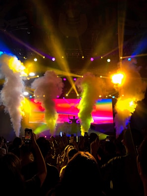 A vibrant concert scene filled with colorful lights and smoke effects. The silhouette of a DJ can be seen on a stage, surrounded by beams of yellow, pink, and blue lights. An energetic crowd raises their hands and phones to capture the experience, contributing to a lively atmosphere.