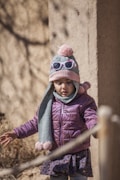 A young child wearing a purple jacket, pink hat with a pom-pom, and a pair of sunglasses on the hat. The child also has a scarf with pom-poms and stands outdoors against a textured wall, partially shaded by natural light.