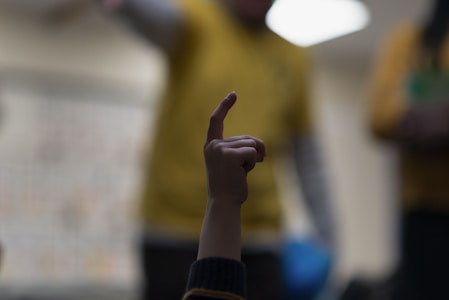 A hand with an index finger pointing upwards is prominently featured in the foreground. The background is blurred, showing figures wearing yellow clothing, suggesting an indoor setting, possibly a classroom or training session.