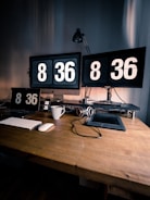A workspace featuring two large monitors each displaying the time 8:36, accompanied by a matching flip clock on a tablet, phone, and digital clock. A keyboard and mouse are placed on the wooden desk, alongside a stylus and a pair of headphones resting on a drawing tablet. A striped coffee mug adds a touch of personal detail to the setup, and an adjustable lamp stands behind the monitors, casting soft light on the scene.