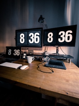 A workspace featuring two large monitors each displaying the time 8:36, accompanied by a matching flip clock on a tablet, phone, and digital clock. A keyboard and mouse are placed on the wooden desk, alongside a stylus and a pair of headphones resting on a drawing tablet. A striped coffee mug adds a touch of personal detail to the setup, and an adjustable lamp stands behind the monitors, casting soft light on the scene.