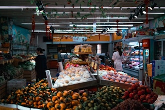 A vibrant indoor market setting with a variety of fresh fruits and vegetables displayed on tables. Different types of produce, such as oranges, apples, and greens, are neatly arranged. People are seen shopping, with a woman in a white coat examining the items. The market is well-lit with ceiling lighting and decorated with hanging ornaments.