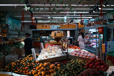 A vibrant indoor market setting with a variety of fresh fruits and vegetables displayed on tables. Different types of produce, such as oranges, apples, and greens, are neatly arranged. People are seen shopping, with a woman in a white coat examining the items. The market is well-lit with ceiling lighting and decorated with hanging ornaments.