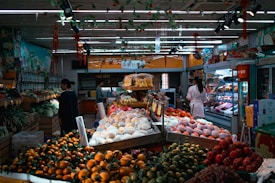 A vibrant indoor market setting with a variety of fresh fruits and vegetables displayed on tables. Different types of produce, such as oranges, apples, and greens, are neatly arranged. People are seen shopping, with a woman in a white coat examining the items. The market is well-lit with ceiling lighting and decorated with hanging ornaments.
