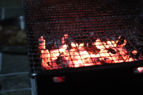 A close-up view of a glowing barbecue grill with orange-red embers, partially covered by a metal grate. The grill is surrounded by darkness, with a faint glimpse of objects, possibly food, on a plate nearby.