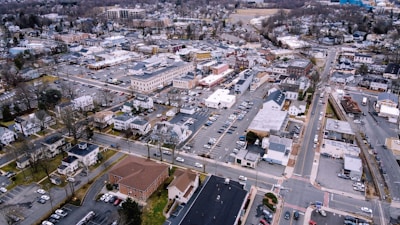 Aerial view of a small town featuring a mix of commercial and residential buildings. The scene includes various cars parked in lots, streets lined with trees, and a variety of rooftops. The town's layout is organized with clearly defined roads and blocks, surrounded by more densely forested areas on the outskirts.