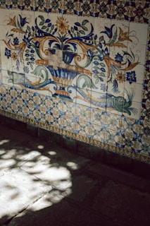 Intricate decorative tiles feature floral and botanical patterns in blue, green, and brown hues. The ornate design includes leaves, flowers, and swirling motifs, set against a backdrop of smaller pattern tiles. Sunlight casts soft, dappled shadows on the tiled floor below.