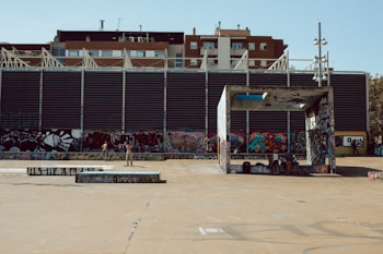 An urban scene featuring a skate park with a large building in the background covered in graffiti art. A few people are visible, with one person skateboarding and others gathered nearby, engaging in conversation. The area is open and expansive with a concrete ground.