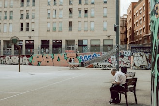 A quiet urban street basketball court with graffiti-covered walls and a solitary elderly man sitting on a bench. The scene is framed by tall residential buildings, and the atmosphere is calm and slightly melancholic.