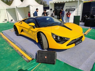A sleek yellow sports car is displayed on a raised platform with a sign that reads 'ROYAL RIDE'. The setting appears to be an outdoor event or exhibition with several white tents visible in the background. There are a few people standing nearby, engaged in conversation, and some informational signage is visible.