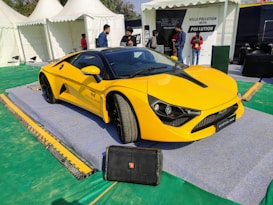 A sleek yellow sports car is displayed on a raised platform with a sign that reads 'ROYAL RIDE'. The setting appears to be an outdoor event or exhibition with several white tents visible in the background. There are a few people standing nearby, engaged in conversation, and some informational signage is visible.