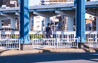 A man in a suit walks across a bridge in an urban area, with elevated roads and various buildings in the background. The scene captures a mix of architectural structures, including blue railings and beams that frame the composition.