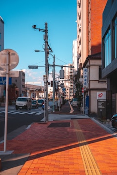 A city street lined with buildings and featuring utility poles with wires. Vehicles, including vans and cars, are visible on the road. A sidewalk with a textured path for visually impaired pedestrians runs alongside the street. The sky is clear and blue, casting shadows across the scene.