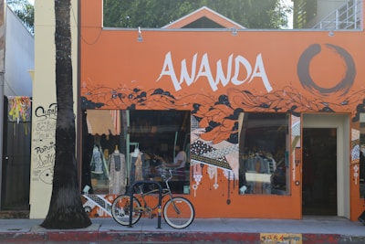 A vibrant storefront with an orange and black mural design featuring abstract patterns and bold lettering 'AWANDA.' A bicycle is parked in front, and mannequins with clothing are visible through the window. A palm tree stands to the left of the entrance, and graffiti art decorates the adjacent wall.