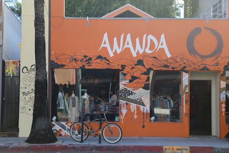 A vibrant storefront with an orange and black mural design featuring abstract patterns and bold lettering 'AWANDA.' A bicycle is parked in front, and mannequins with clothing are visible through the window. A palm tree stands to the left of the entrance, and graffiti art decorates the adjacent wall.