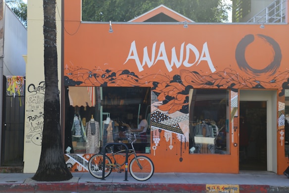 A vibrant storefront with an orange and black mural design featuring abstract patterns and bold lettering 'AWANDA.' A bicycle is parked in front, and mannequins with clothing are visible through the window. A palm tree stands to the left of the entrance, and graffiti art decorates the adjacent wall.