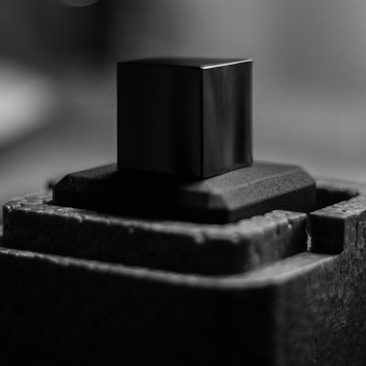 A black and white close-up of a cube resting on a textured surface. The lighting is soft, creating gentle shadows and highlights on the cube's edges. The background is blurred, focusing attention on the cube.