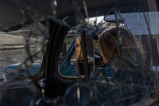 A woman is sitting inside a vehicle, looking at her reflection in the side mirror. The windshield appears cracked, creating an intricate web pattern across the image. The setting seems to be in an outdoor, possibly rural area with another old vehicle visible through the window.