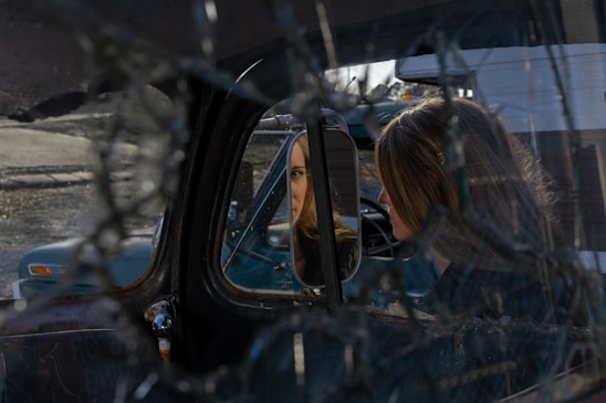 A woman is sitting inside a vehicle, looking at her reflection in the side mirror. The windshield appears cracked, creating an intricate web pattern across the image. The setting seems to be in an outdoor, possibly rural area with another old vehicle visible through the window.