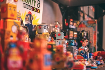 A collection of colorful vintage toy robots displayed together. These toys include various designs with metallic features and bright colors, creating a nostalgic atmosphere. Some robots are standing upright, while others are lying on their sides. There are also other toys and items in the background, adding to the busy and playful setup.