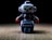 A small, toy robot stands on a flat surface with a dark background. The robot is made of plastic, featuring a predominantly blue-gray body with red accents on its arms and head. Its eyes are large and white, giving it a cartoonish appearance.