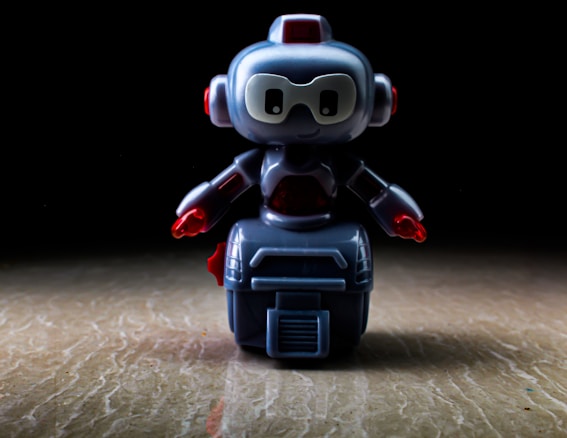 A small, toy robot stands on a flat surface with a dark background. The robot is made of plastic, featuring a predominantly blue-gray body with red accents on its arms and head. Its eyes are large and white, giving it a cartoonish appearance.