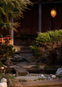 A serene garden setting features a small stone path leading to a wooden deck. A hanging lantern softly illuminates the scene, casting a warm glow. Evergreen shrubs and neatly arranged rocks border the area, contributing to a tranquil ambiance.