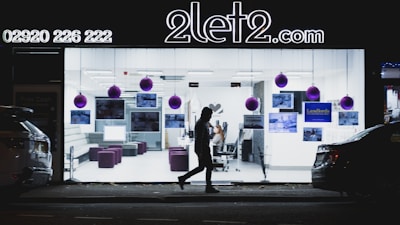 A brightly lit office space is visible through a large glass window. The office interior includes several chairs, a couch, and wall-mounted images. Purple ornaments hang from the ceiling. A silhouette of a person walks in front of the window, and two cars are parked on either side of the scene.
